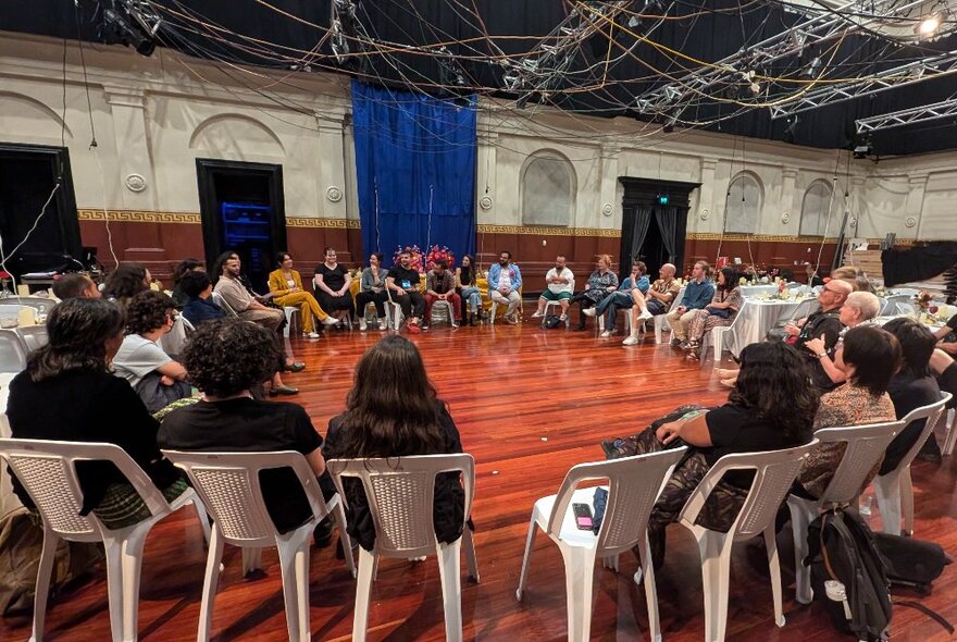 People seated on white plastic chairs that form a large circle inside the main hall in a large arts venue.