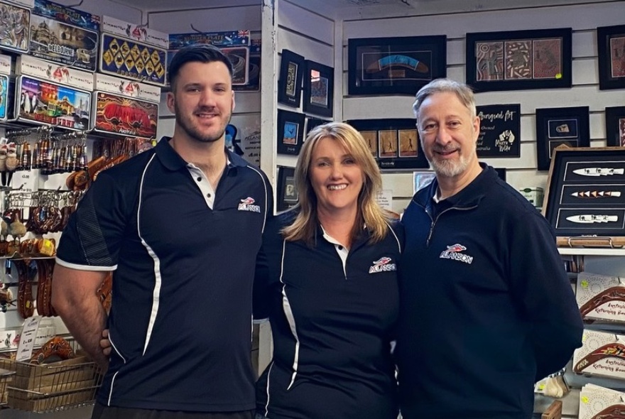 Three store workers wearing navy uniforms, standing in front of shelves of jigsaws, souvenirs and boomerangs.