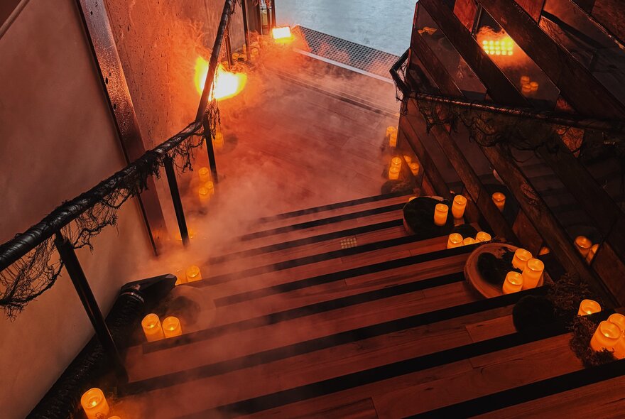 A dimly lit staircase lined with glowing orange pillar candles along the steps and handrails, creating a moody, atmospheric entrance.