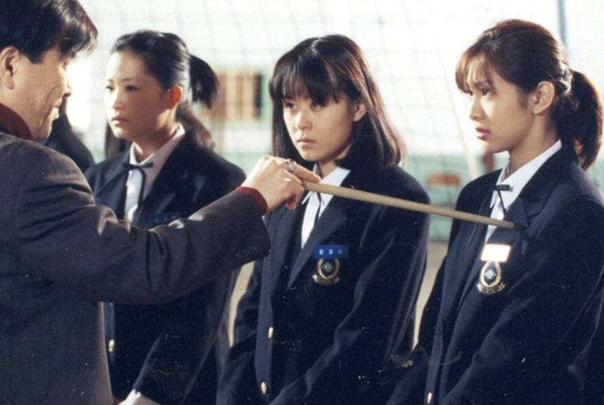 A still from a Korean horror film showing three school girls and a male school teacher pointing at one of them with a wooden pointer.