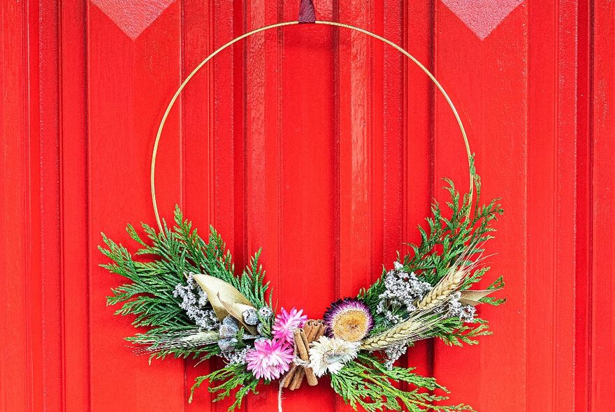 A round Christmas wreath of fresh native flowers and foliage, hanging against a red curtain.