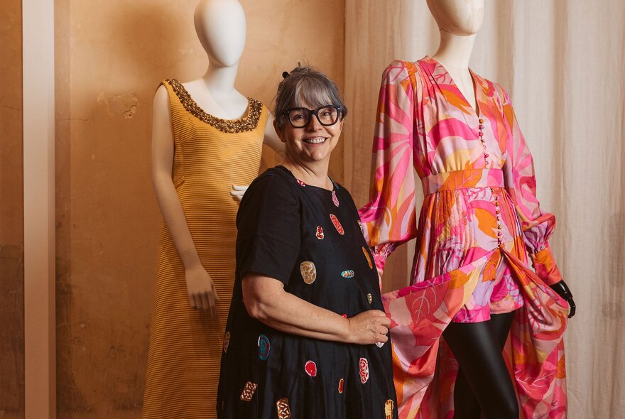 Woman wearing a dark patterned dress, smiling in front of two mannequins wearing brightly coloured gowns.