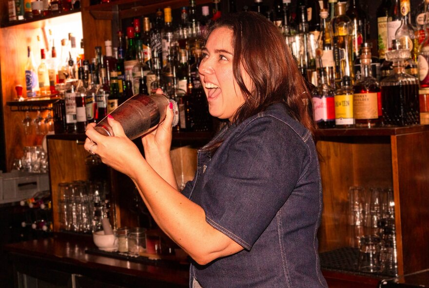 A person smiling while shaking a cocktail in a metal cocktail shaker, behind a bar