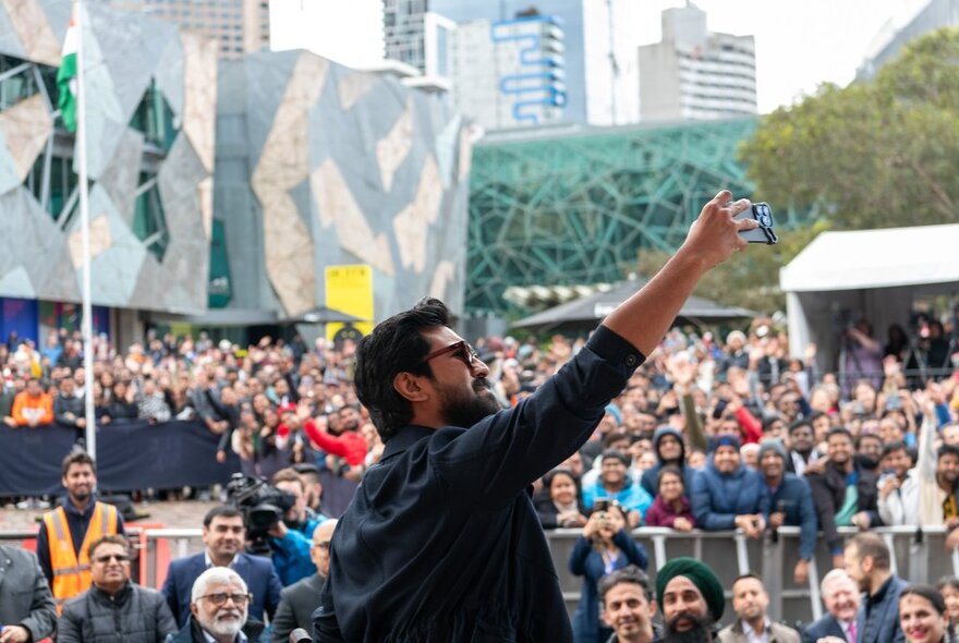 A bearded man in sunglasses takes a selfie with a huge crowd behind him in Federation Square. 
