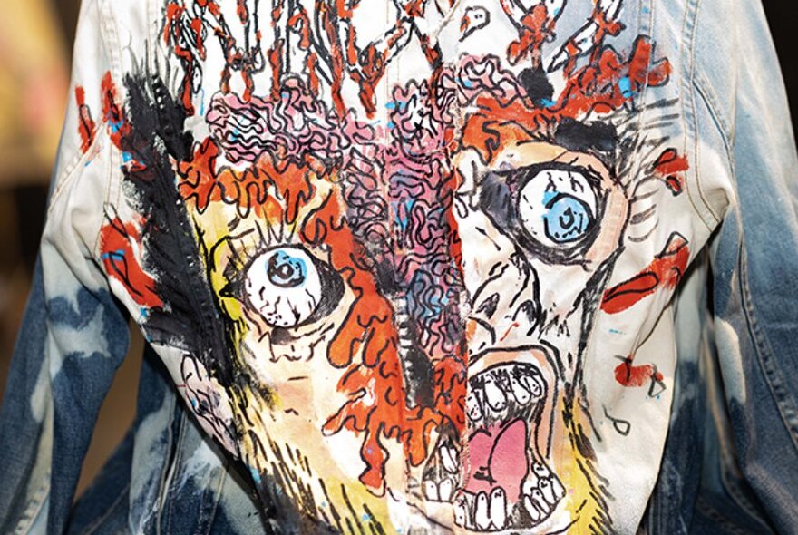 The painted back of a denim jacket, featuring a skeletal face with two eyes, an open mouth and orange and pink brains.