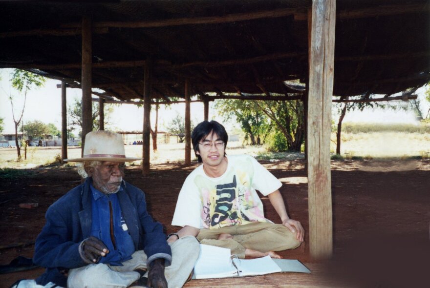 A Japanese man seated on the ground next to an Aboriginal Elder.