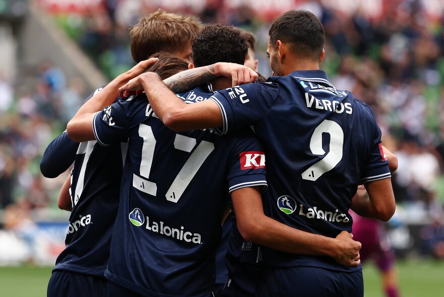 Behind view of three soccer players on a field with their arms around each other. 