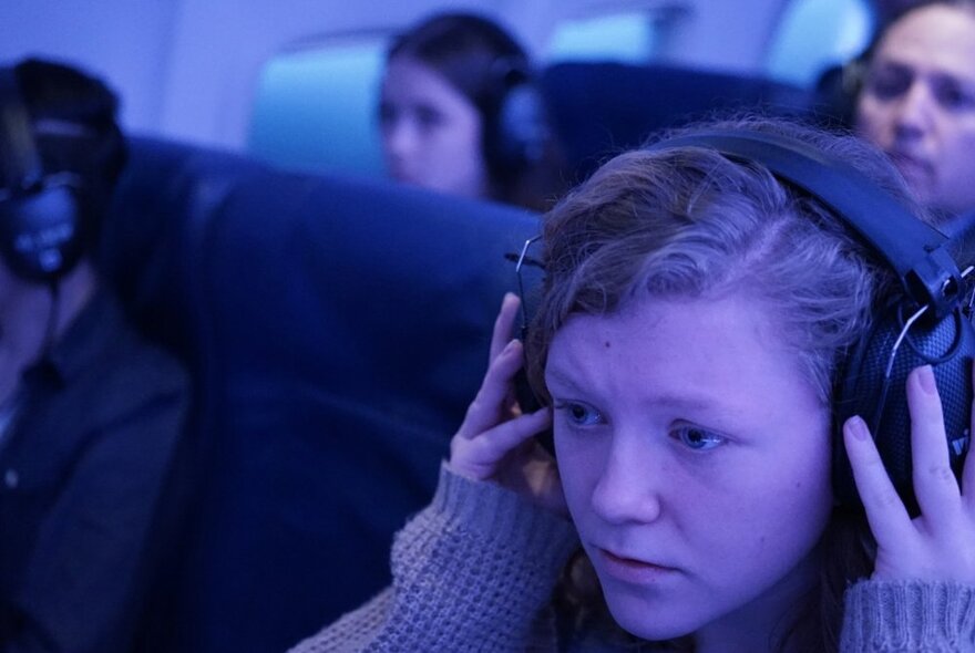 A person seated in an airline seat, with their hands on a pair of headphones they are wearing on their head, a pensive expression on their face.