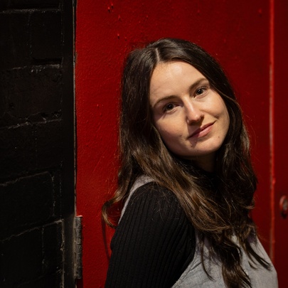 Woman with long dark hair, standing in front of red background.