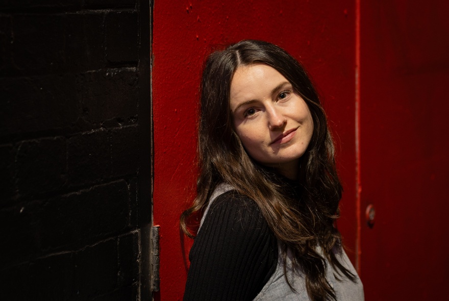 Woman with long dark hair, standing in front of red background.