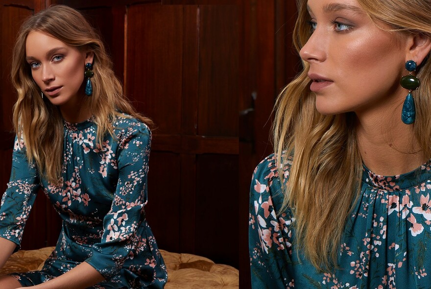 Model wearing a blue floral dress seated by a mirror, her reflection gazing at the camera.