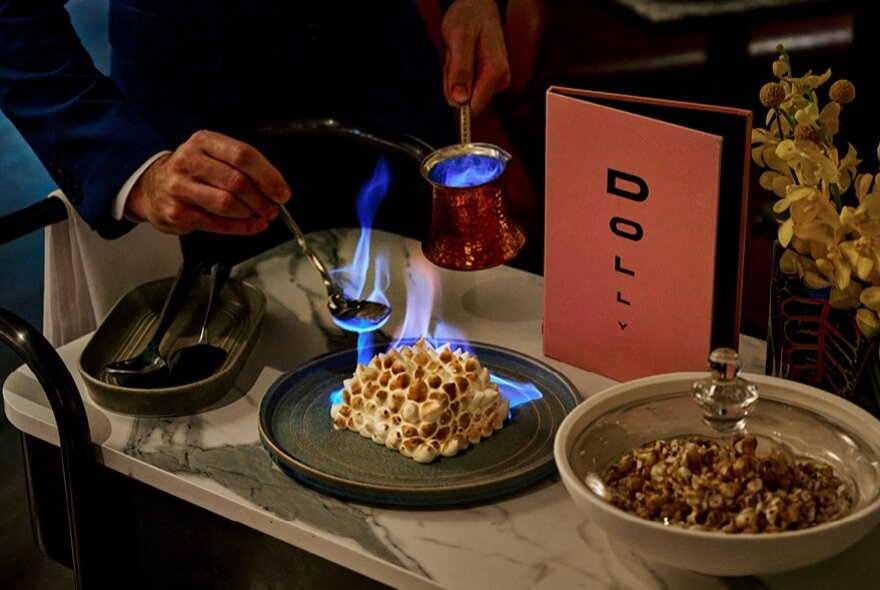 A waiter in black pouring flaming liqueur onto a small bombe alaska on a marble table alongside a folded menu and a glass covered dish with granola.