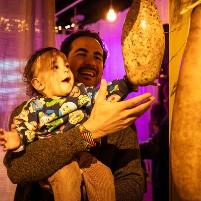 A smiling man holds a cheerful toddler in his arms as the child reaches toward a large, textured, teardrop-shaped gourd hanging from the ceiling; part of an interactive installation at ArtPlay.