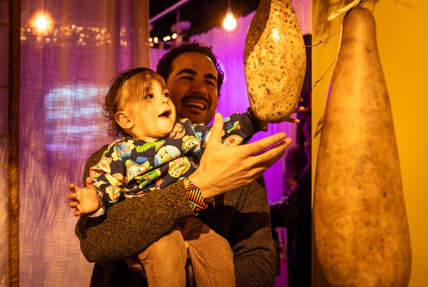 A smiling man holds a cheerful toddler in his arms as the child reaches toward a large, textured, teardrop-shaped gourd hanging from the ceiling; part of an interactive installation at ArtPlay.