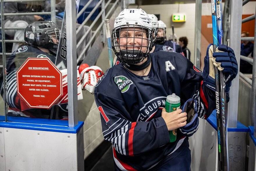Female ice hockey player holding a water bottle, smiling through her helmet guard, standing in arena walkway.