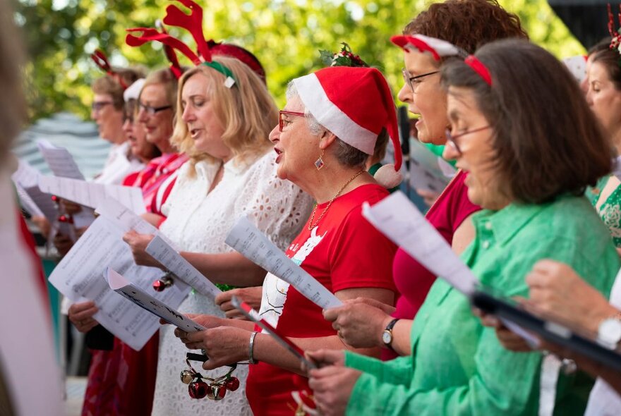 A row of amateur singers in a choir, wearing Christmas colours of red and green, some wearing red hats, holding sheet music and singing together.