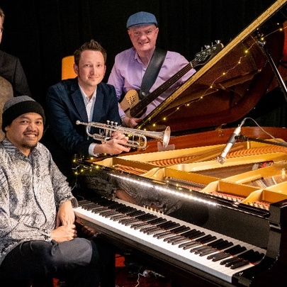 Jazz musicians gathered around a grand piano, with pianist seated at the piano, other musicians holding instruments.