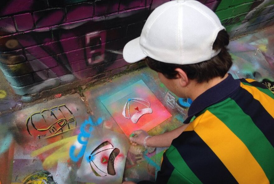 A young person wearing a baseball cap using a spray can to make stencil art, outside in a laneway.