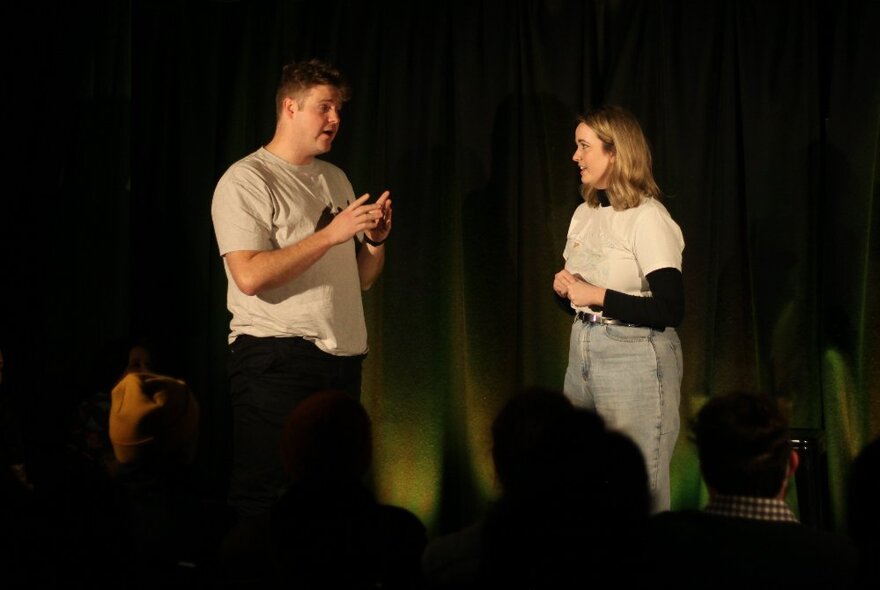 Two performers on a stage in front of a curtain, interacting with one another and improvising a scene in front of an audience.