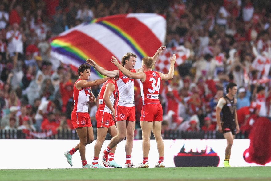Sydney Swans players huddle together during a match, a large flag in the background. 