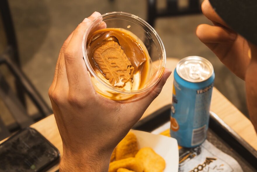 A person holding a clear plastic cup filled with a creamy dessert over a tray at a table in a fast food restaurant.