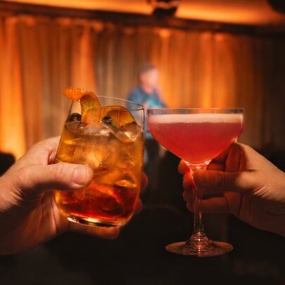 Two hands clinking cocktails in a room with a small stage. 