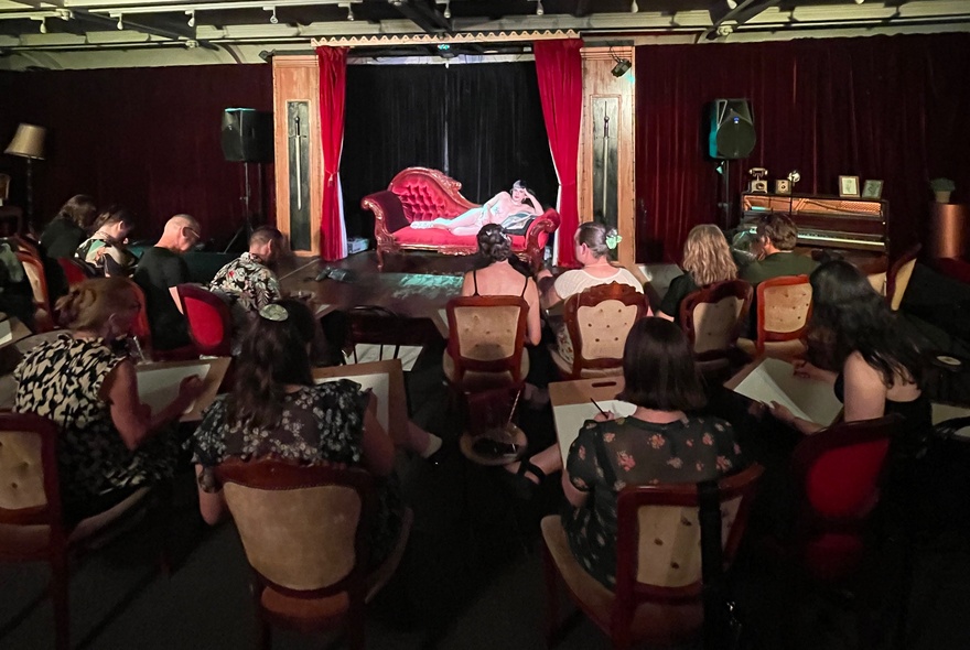 A life drawing class in a small red room, a model reclining a the front with people seated drawing. 