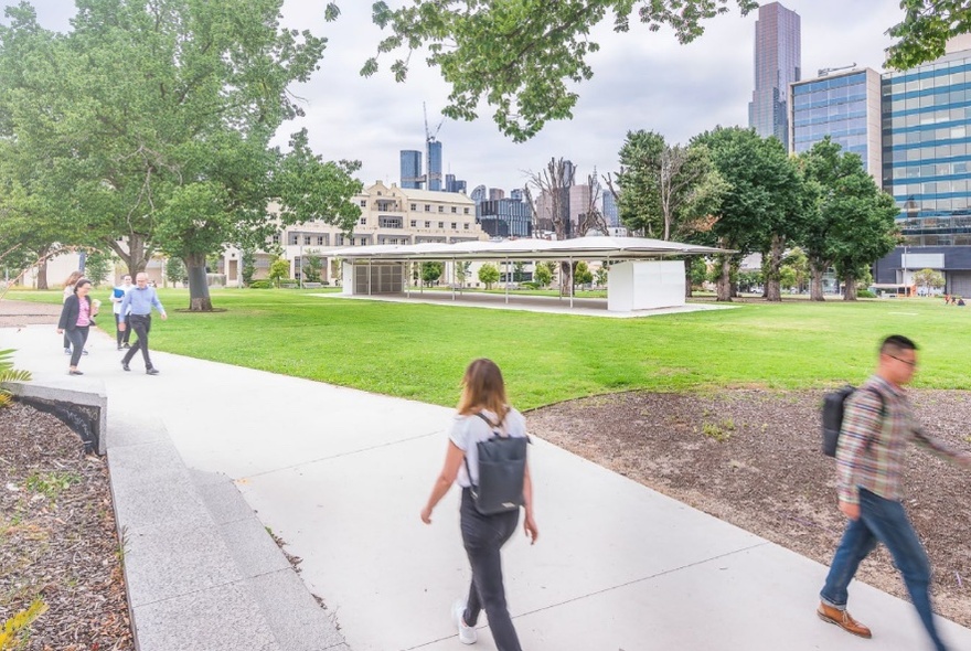 People walking on a concrete footpath past the MPavilion, an open-concept building with a flat roof structure surrounded by grass in front of the Melbourne cityscape.