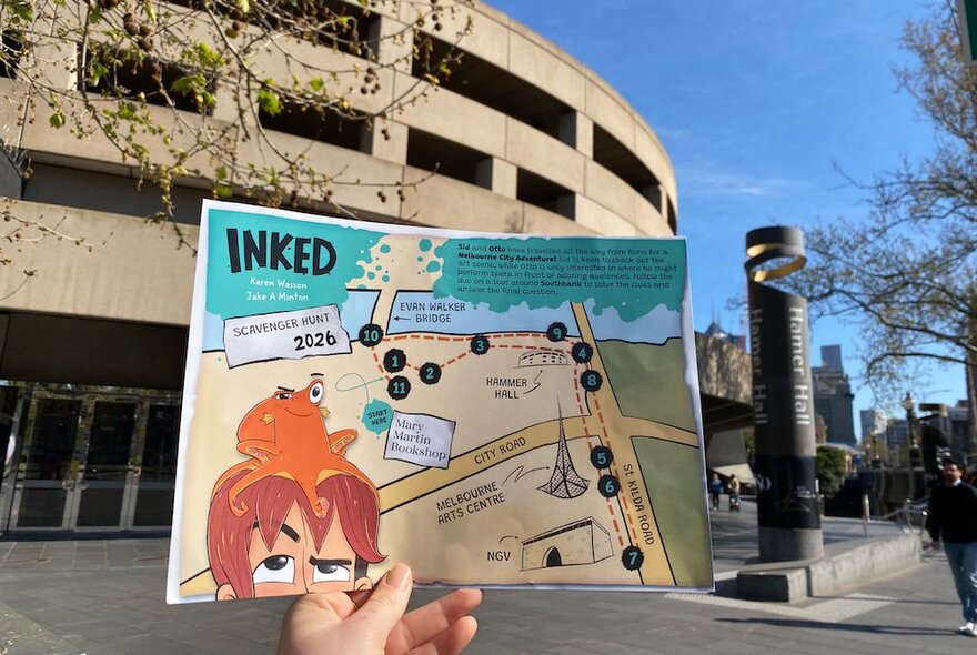 A person holding an 'Inked' scavenger map in front of the Hamer Hall, Arts Centre Melbourne with a blue sky background.