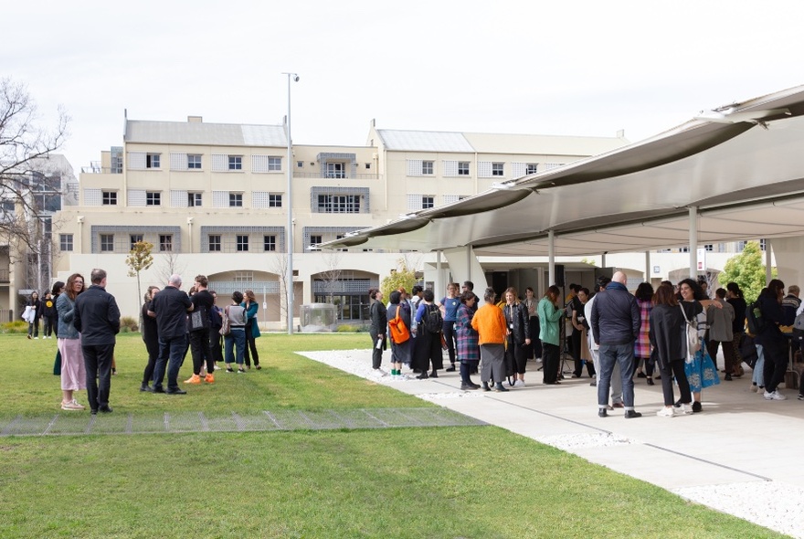 A crowd of people participating in an activity at the MPavilion, an open-concept building with a flat roof structure, surrounded by grass with multi-storey housing behind.