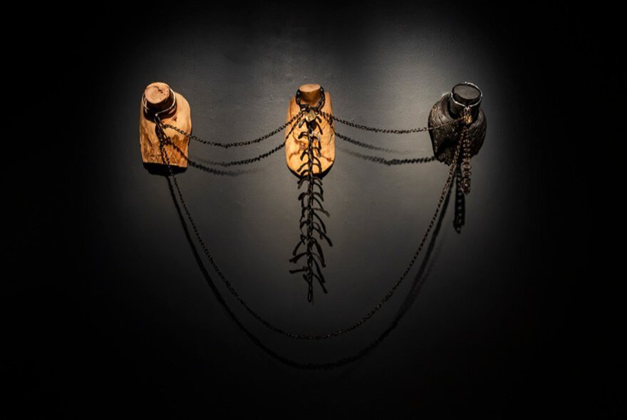 A black wall with light on artwork showing three hooks with wires attached to them.
