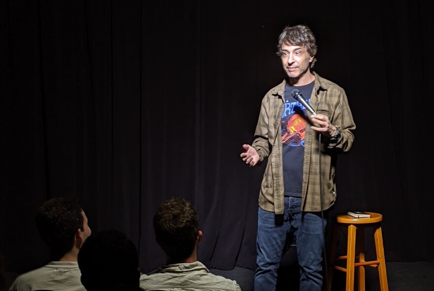 Comedian, Arj Barker, on stage next to a wooden stool, wearing a flannel shirt over a t-shirt and holding a microphone.