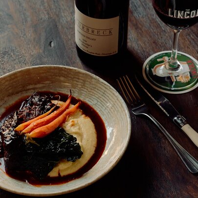 A plate of braised beef and mashed potato on a wooden table alongside a bottle of wine and a filled wine glass.
