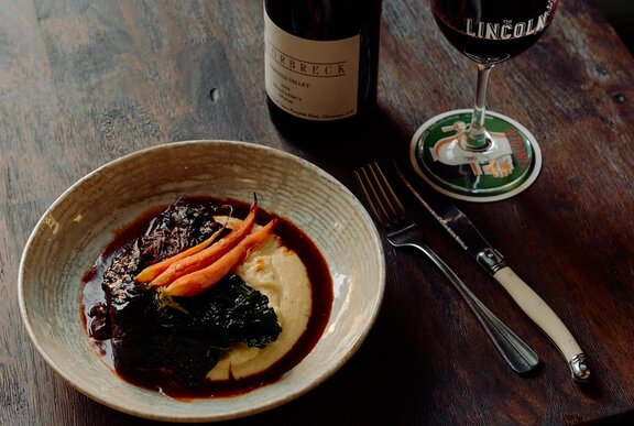 A plate of braised beef and mashed potato on a wooden table alongside a bottle of wine and a filled wine glass.