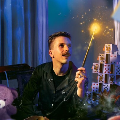 Magician sitting cross legged on a cosy bed in a darkened room, wearing pyjamas and holding a wand, with a tower of cards and a teddy bear as props that surround him.
