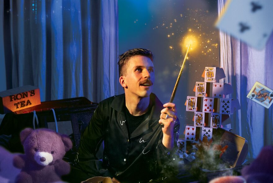 Magician sitting cross legged on a cosy bed in a darkened room, wearing pyjamas and holding a wand, with a tower of cards and a teddy bear as props that surround him.