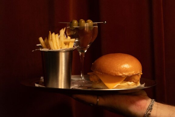 A hand holding a tray that contains a fish burger, a container of fries and a martini.