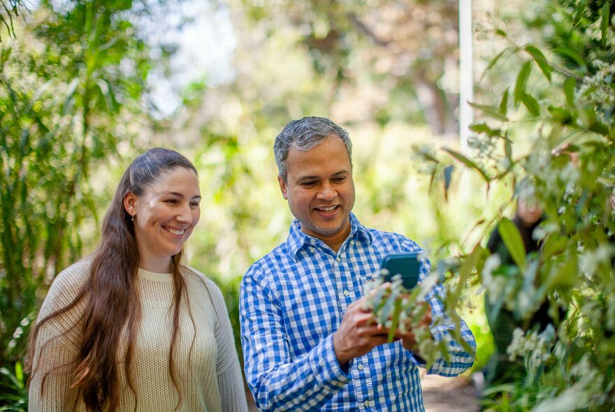 Two people smiling as they walk through a garden, the man looking at his phone. 