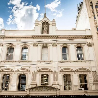 The ornate, light-coloured facade of Melbourne Athenaeum Library, featuring classical architectural details, multiple windows on two main floors, decorative columns, and a central statue niche at the top under a cloudy blue sky.