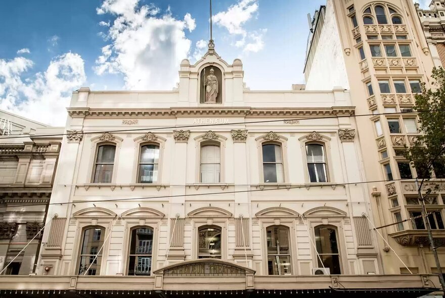 The ornate, light-coloured facade of Melbourne Athenaeum Library, featuring classical architectural details, multiple windows on two main floors, decorative columns, and a central statue niche at the top under a cloudy blue sky.