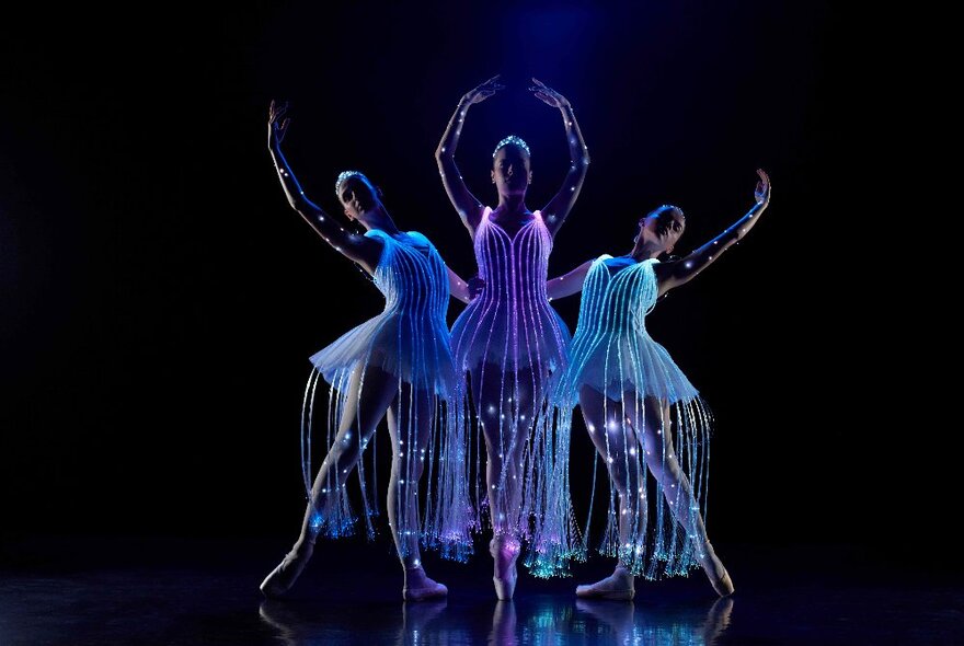 Dark stage with ultraviolet-lit ballet dancers pirouetting as a trio.