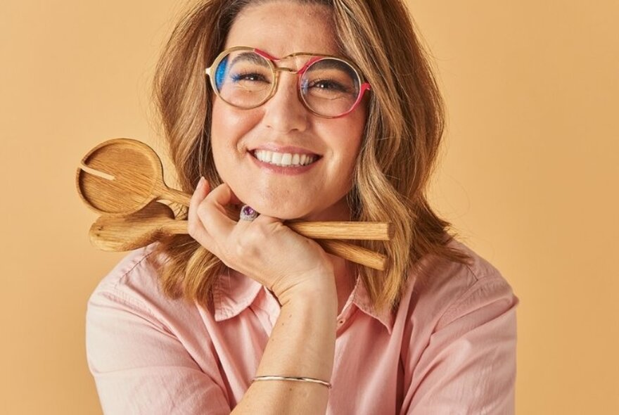Food writer and author Alice Zaslavsky, smiling, wearing glasses and holding wooden salad servers in her hands that rest under her chin.