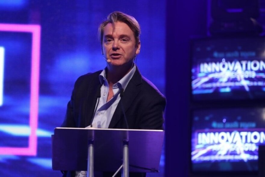 Rockstar global economist, David McWilliams, standing in front of a podium on a brightly lit stage with blue lights and screens behind him.