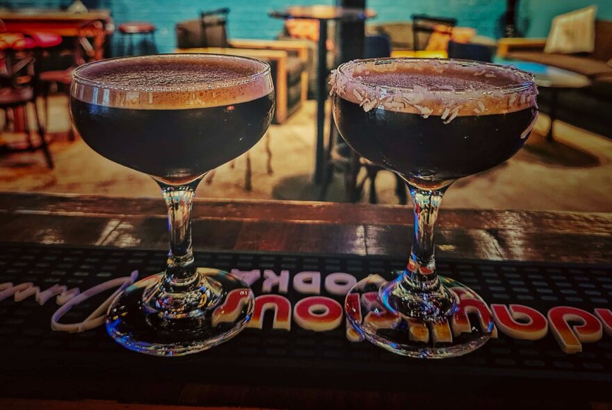 Two espresso martinis on a bar counter, with tables and chairs in the background.