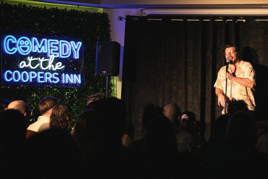Comedian holding a microphone on a small curtained stage, a neon sign of the name of the club room visible to his right that reads COMEDY at the COOPERS INN.