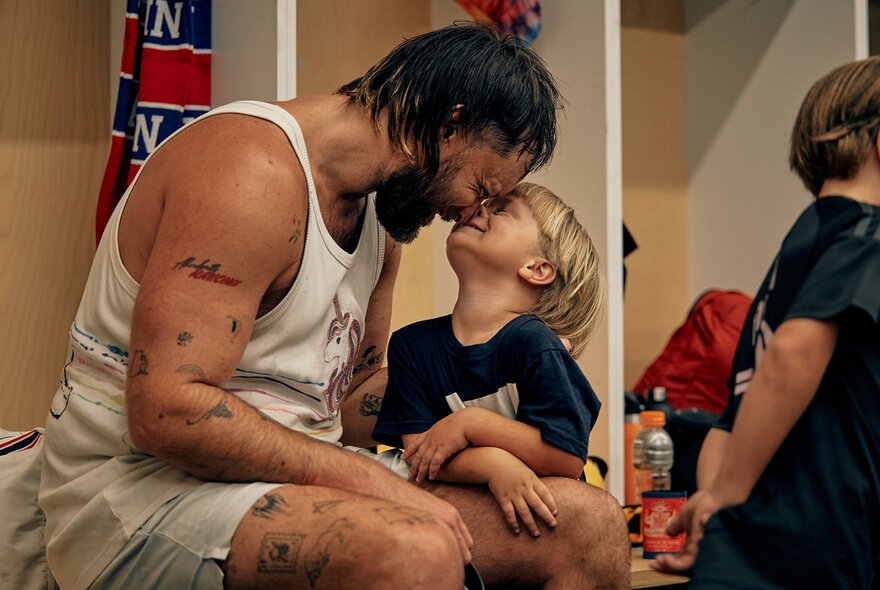 A candid, intimate image inside a sports locker room shows a bearded man in a white tank top sitting down, leaning forward to gently press his forehead against the forehead of a smiling young blonde boy sitting on his lap, his son.
