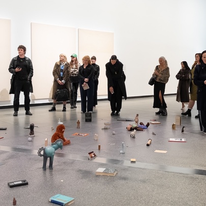 People gathered in a gallery space installation that consists of found objects, photographs, and ephemera scattered across the floor, listening to a person hosting a talk.