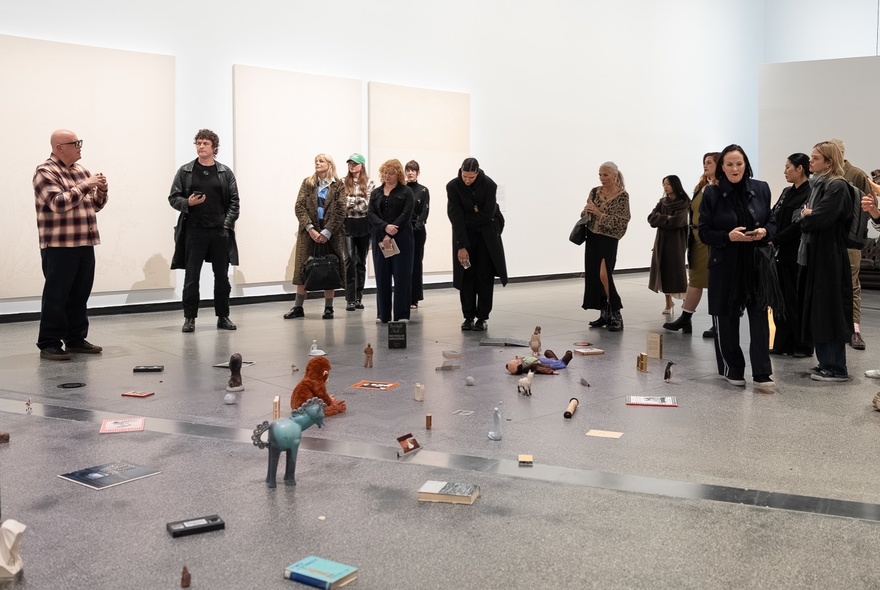 People gathered in a gallery space installation that consists of found objects, photographs, and ephemera scattered across the floor, listening to a person hosting a talk.