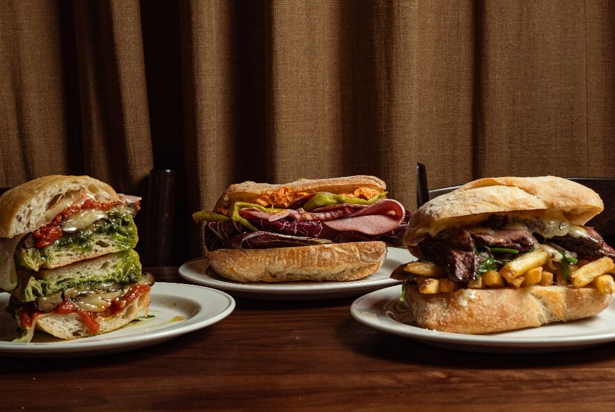 Three enormous sandwiches with different fillings displayed on white plates set on a wooden table in front of brown curtain.
