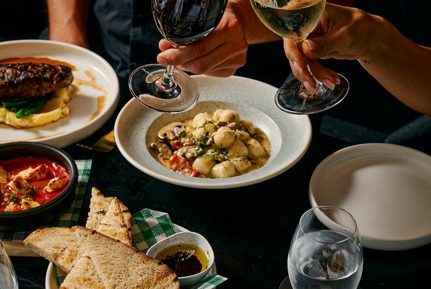 A selection of Italian food, with a gnocci dish, steak and mash and panini bread, with two hands holding wine glasses.